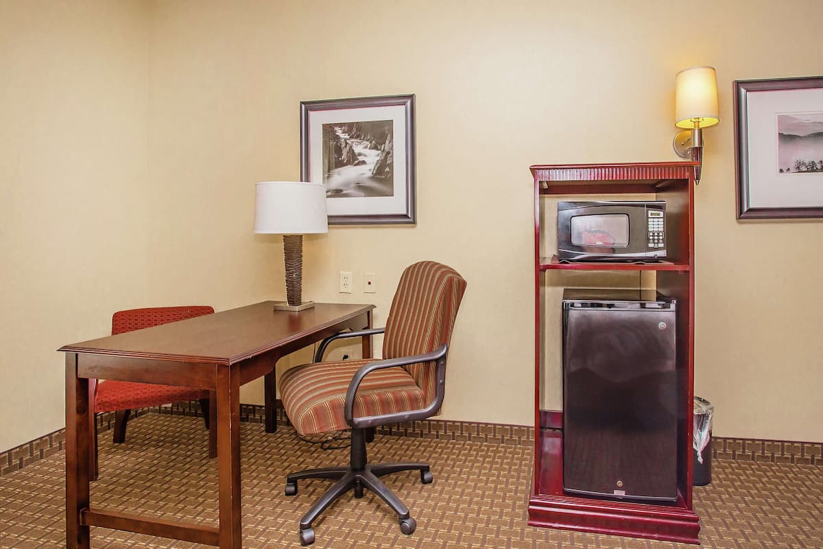 A hotel room with a wooden desk, a red chair, a microwave, a refrigerator, a lamp, and two framed pictures on the wall.