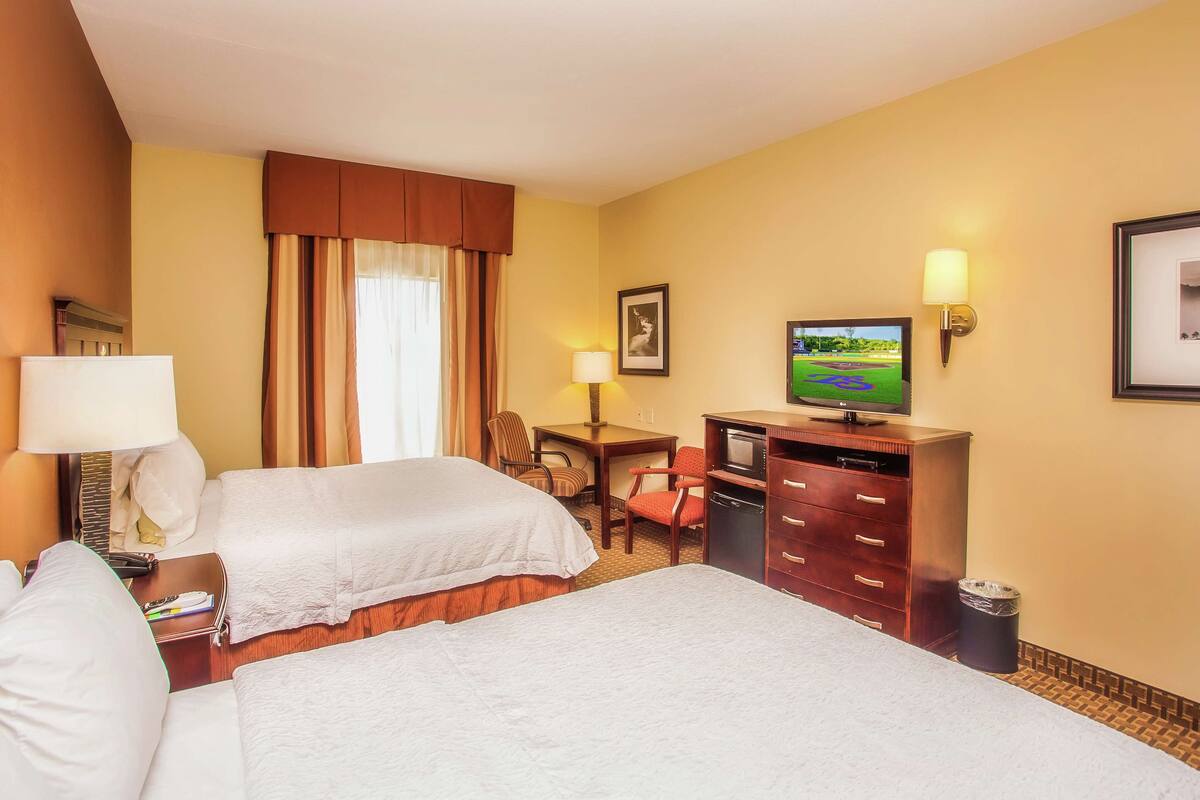 A hotel room with two beds, a desk, a television, and a trash can.
