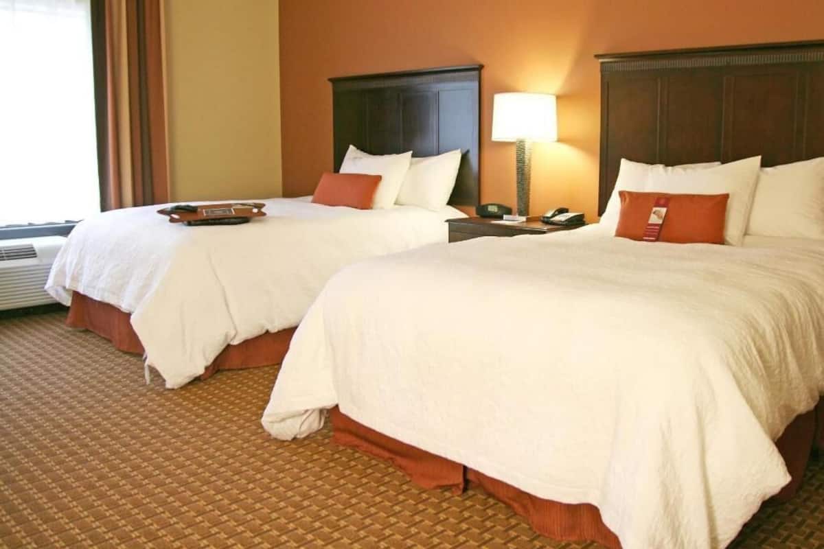 A hotel room with two beds, a nightstand with a lamp, and a window with curtains.