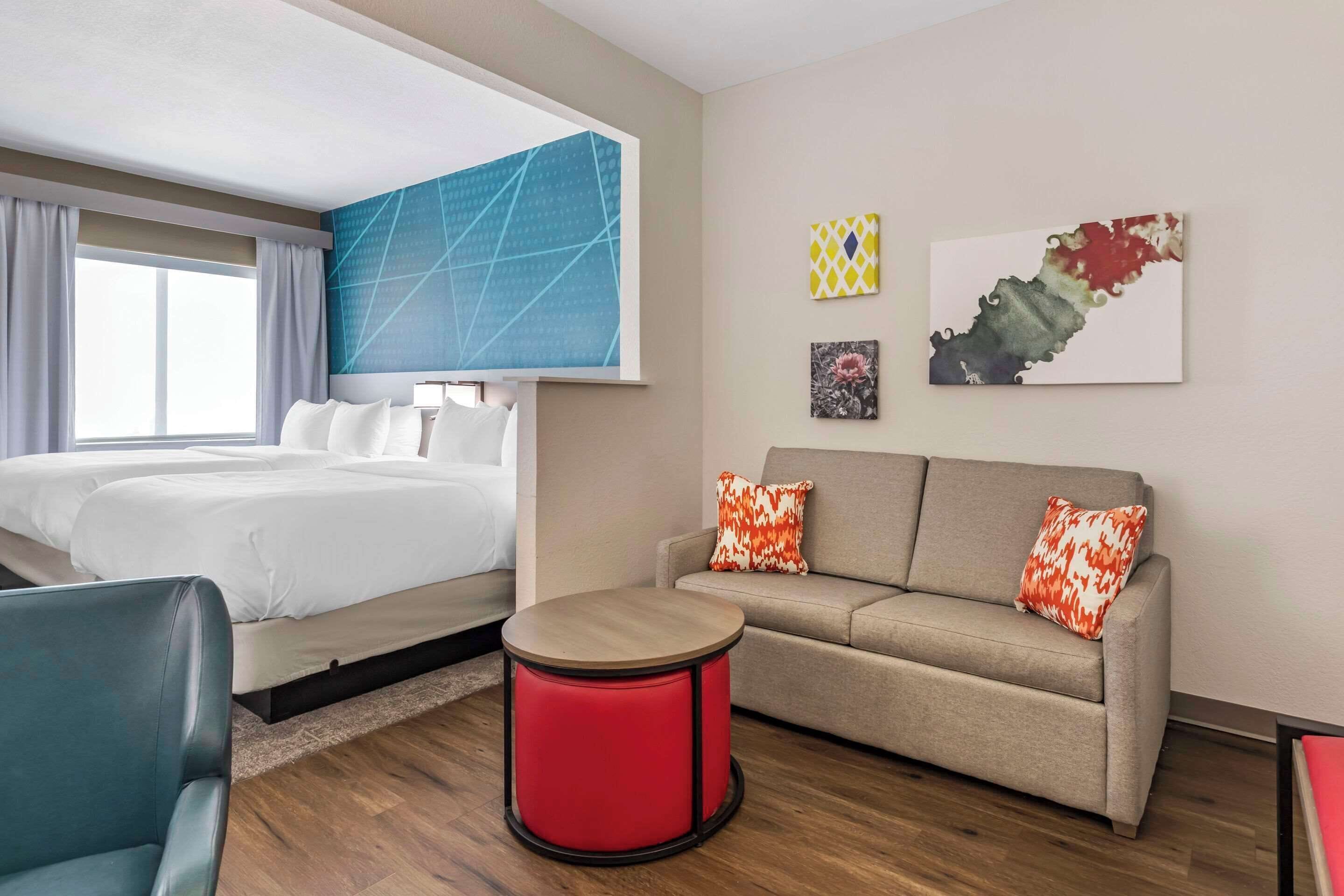 Una camera d'albergo con un letto, una poltrona, un poggiapiedi rosso e opere d'arte alle pareti.