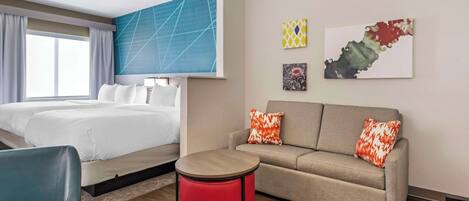 A hotel room with a bed, a sofa, a red ottoman, and artwork on the walls.