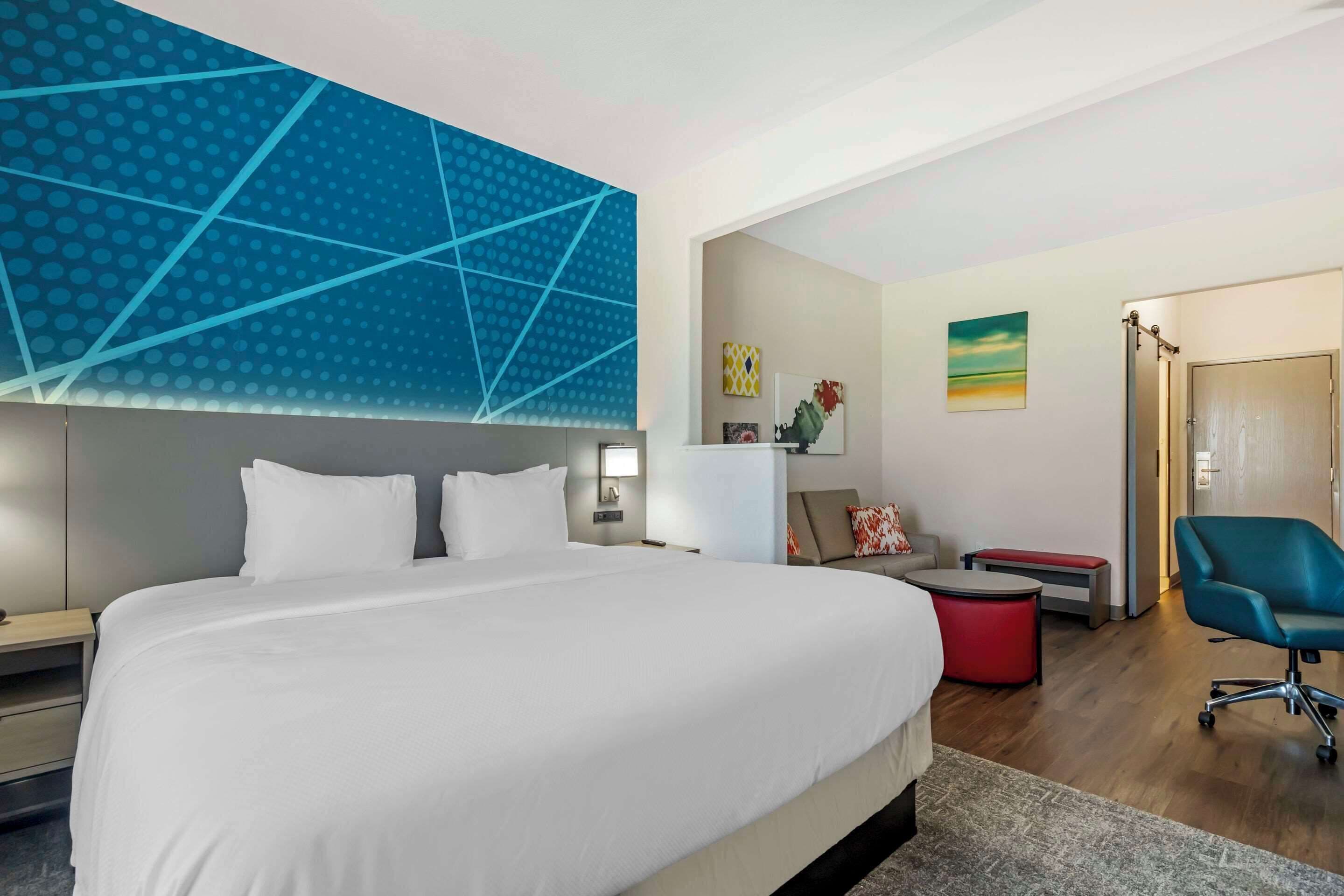 Une chambre d’hôtel moderne avec un grand lit, un bureau, une chaise et un petit pouf rouge.