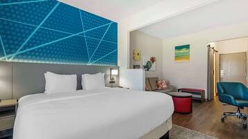 A modern hotel room with a large bed, a desk, a chair, and a small red ottoman.
