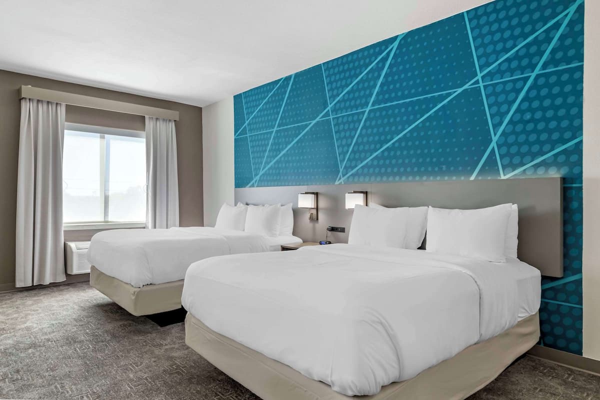 Ein Hotelzimmer mit zwei Betten, einem großen Fenster mit Vorhängen, einer Wand mit geometrischem Muster und einem an der Wand befestigten Nachttisch mit Lampe.