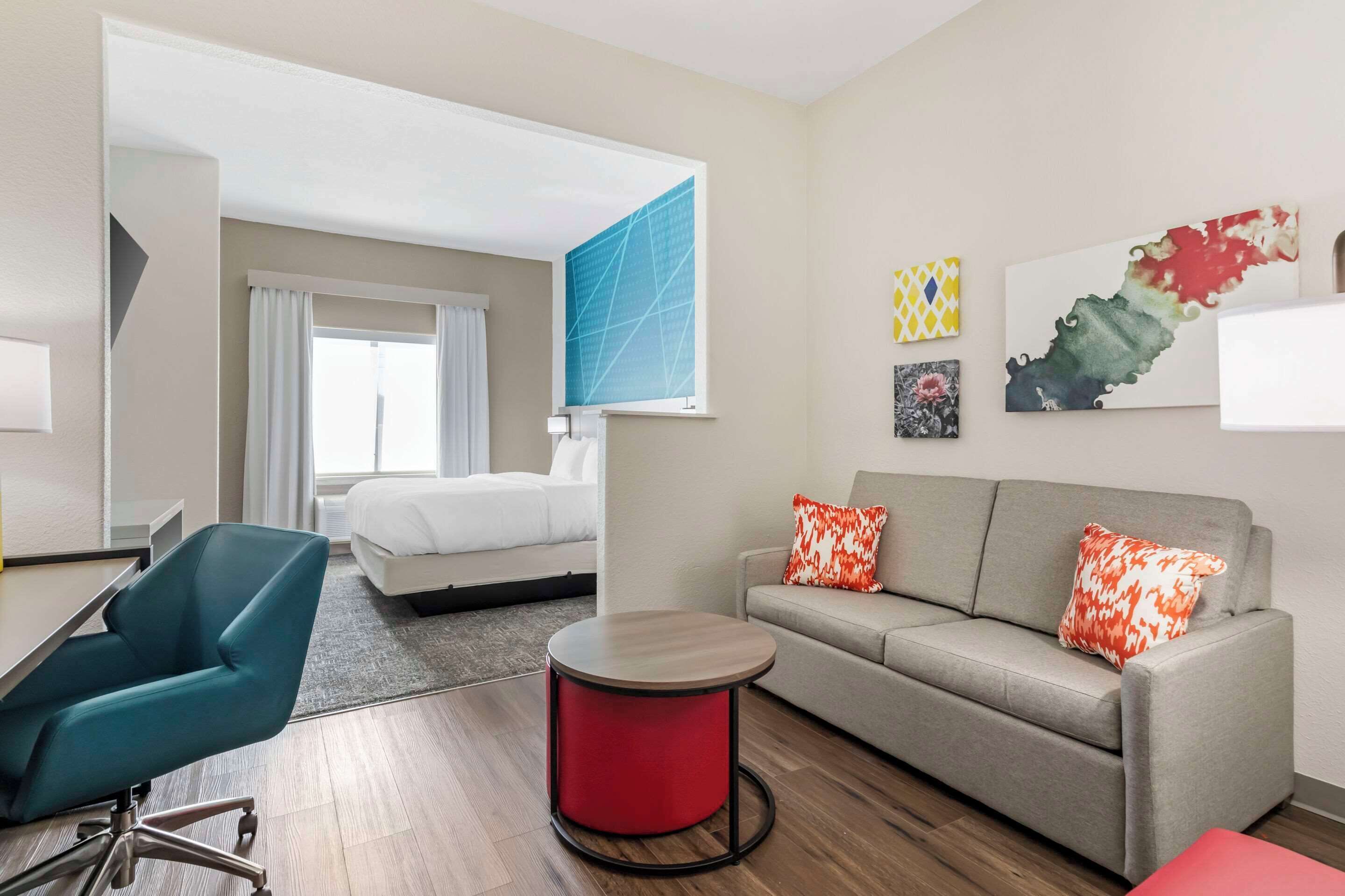 A hotel room with a bed, a sofa, a desk, and a chair. There are artworks on the wall and a window with curtains.