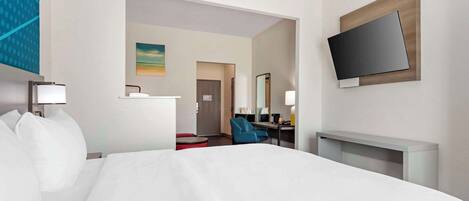 Habitación de hotel con una cama grande, un televisor de pantalla plana montado en la pared, un escritorio con una silla y un pequeño rincón de descanso con un reposapiés rojo.