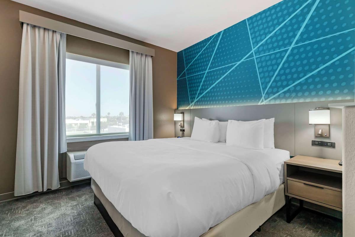 Ein Hotelzimmer mit einem großen Bett, Blick auf die Stadt, einer modernen blauen Akzentwand und einem Nachttisch mit Lampe.