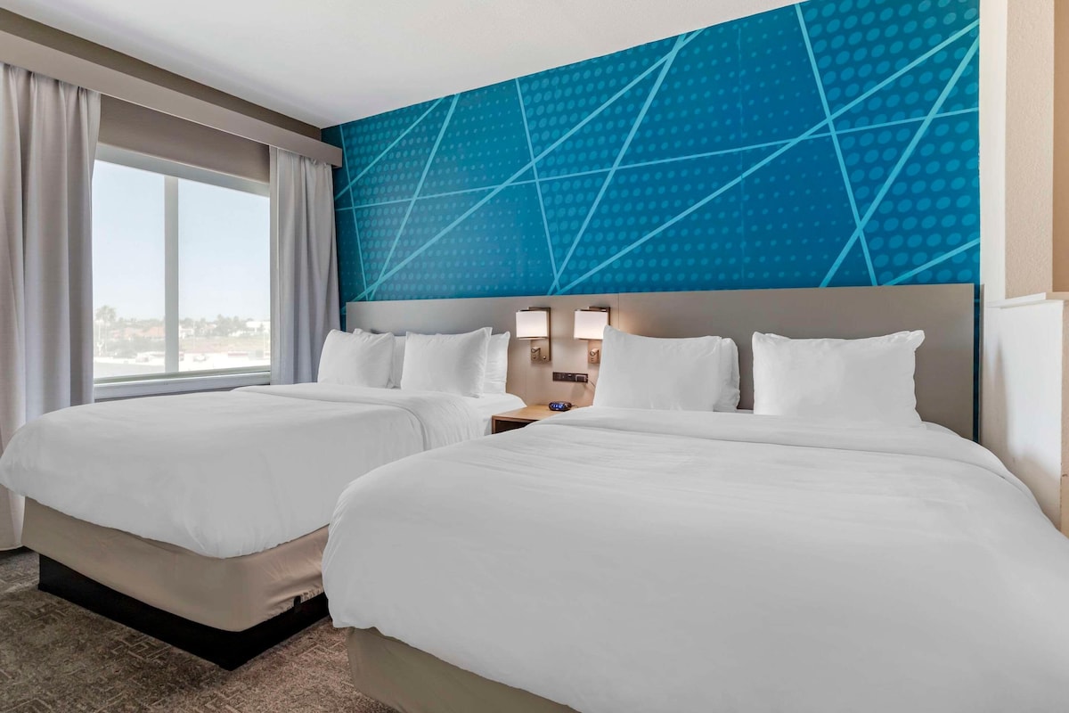 A hotel room with two beds, a large window, and a blue geometric wall design.