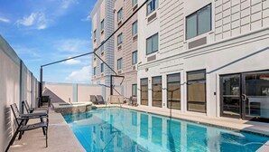 En swimmingpool med solstolar och en solterrass, omgiven av en hög byggnad med stora fönster och en modern design.