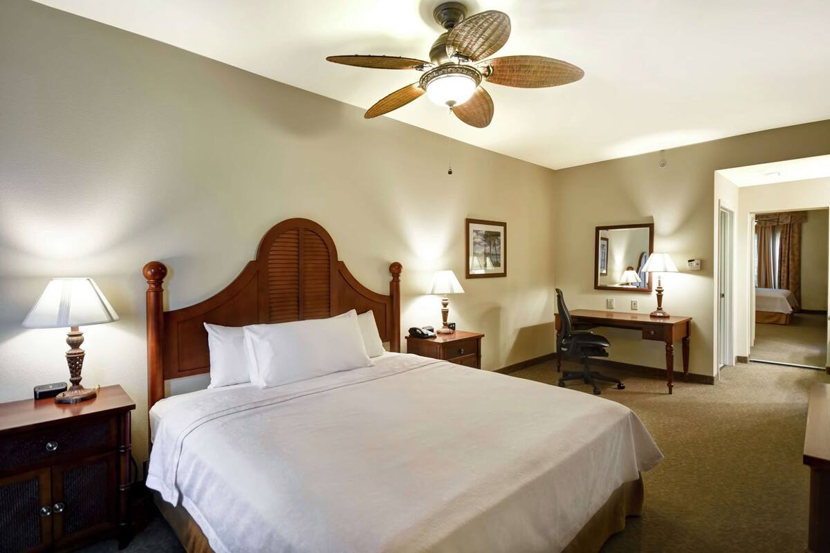 A hotel room with a large bed, a desk, a chair, a ceiling fan, and a mirror.