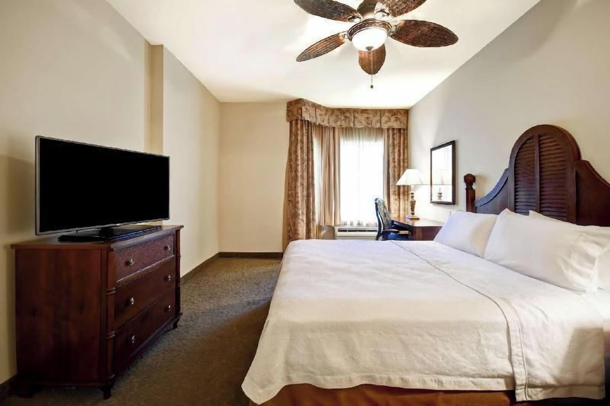 Una habitación de hotel con cama, televisor en una cómoda, ventilador de techo y ventana con cortinas.
