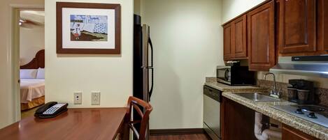 A compact kitchen with wooden cabinets, a refrigerator, microwave, and a coffee maker. A dining table with chairs is positioned near the kitchen, and a framed picture hangs on the wall.