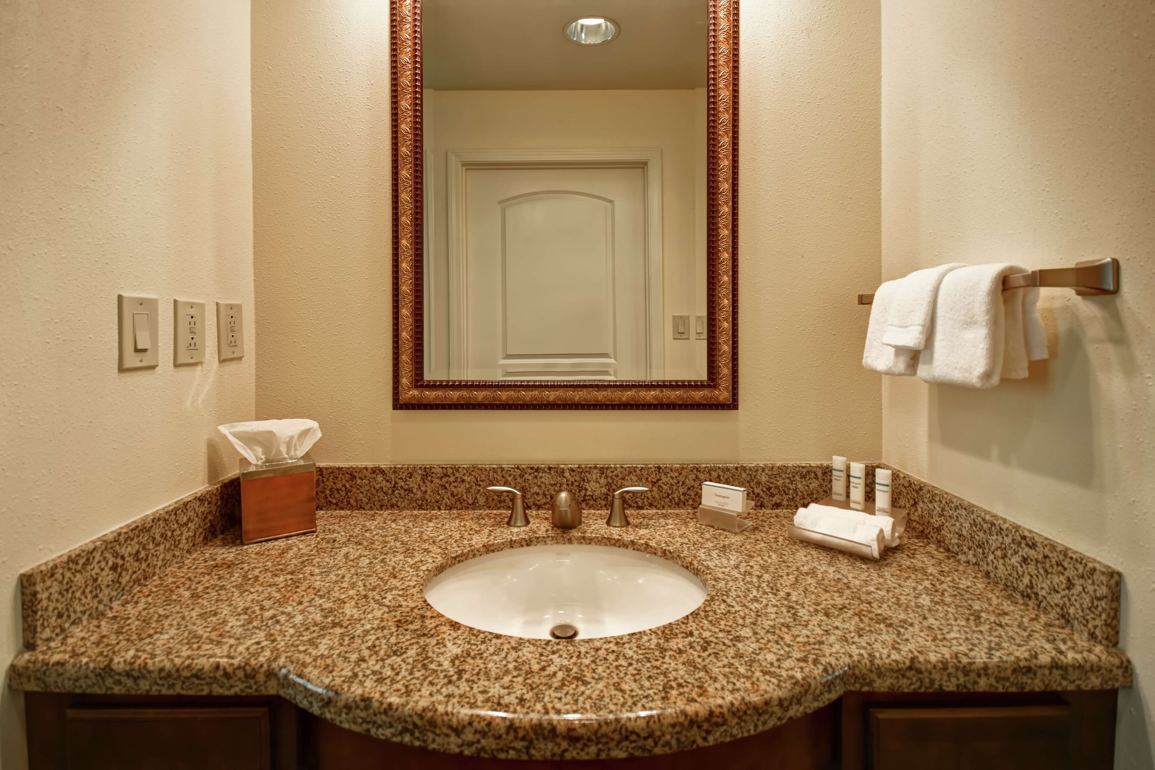 A bathroom with a granite countertop sink, a mirror with a decorative frame, a towel rack, and a tissue box.