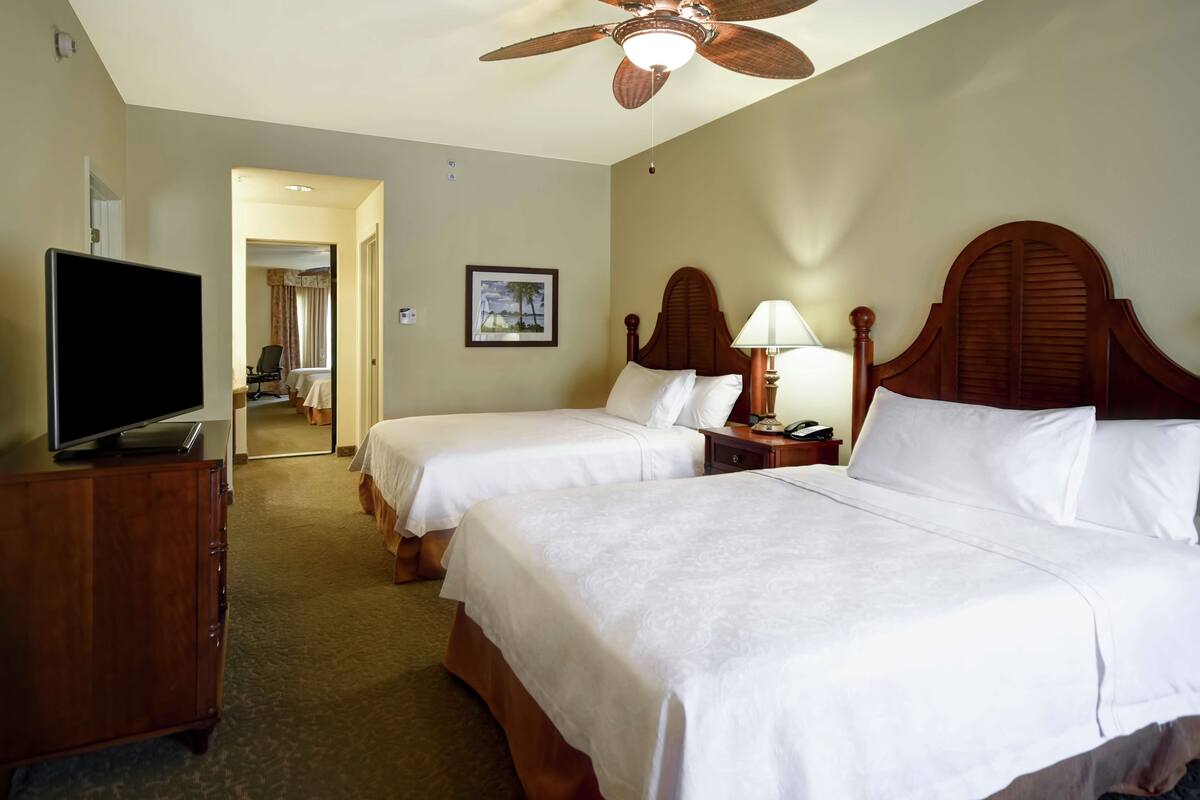 A hotel room with two beds, a television on a dresser, a ceiling fan, and a framed picture on the wall.