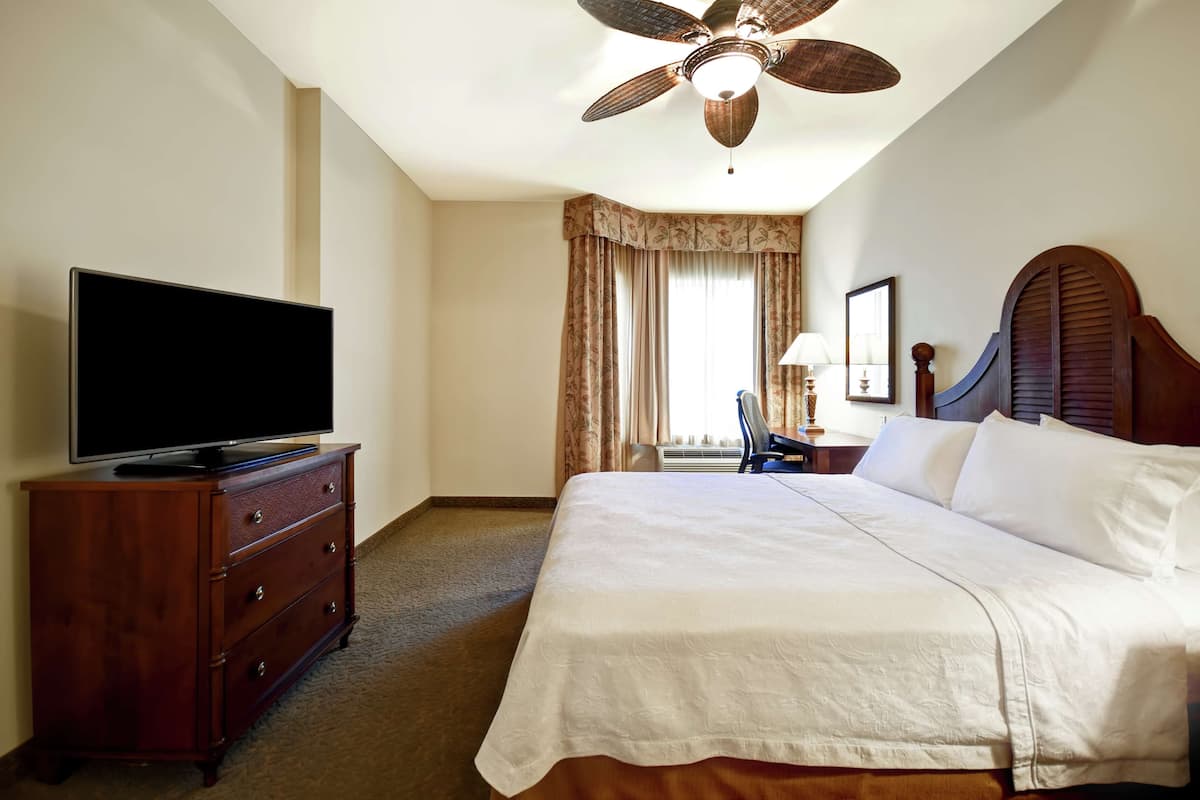 A hotel room with a bed, a television on a dresser, a ceiling fan, and a window with curtains.