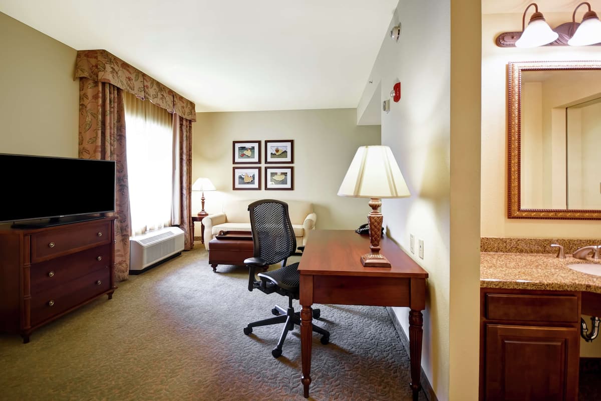 A hotel room with a flat-screen TV, a wooden desk with a lamp, a black office chair, a sofa, and a large mirror.