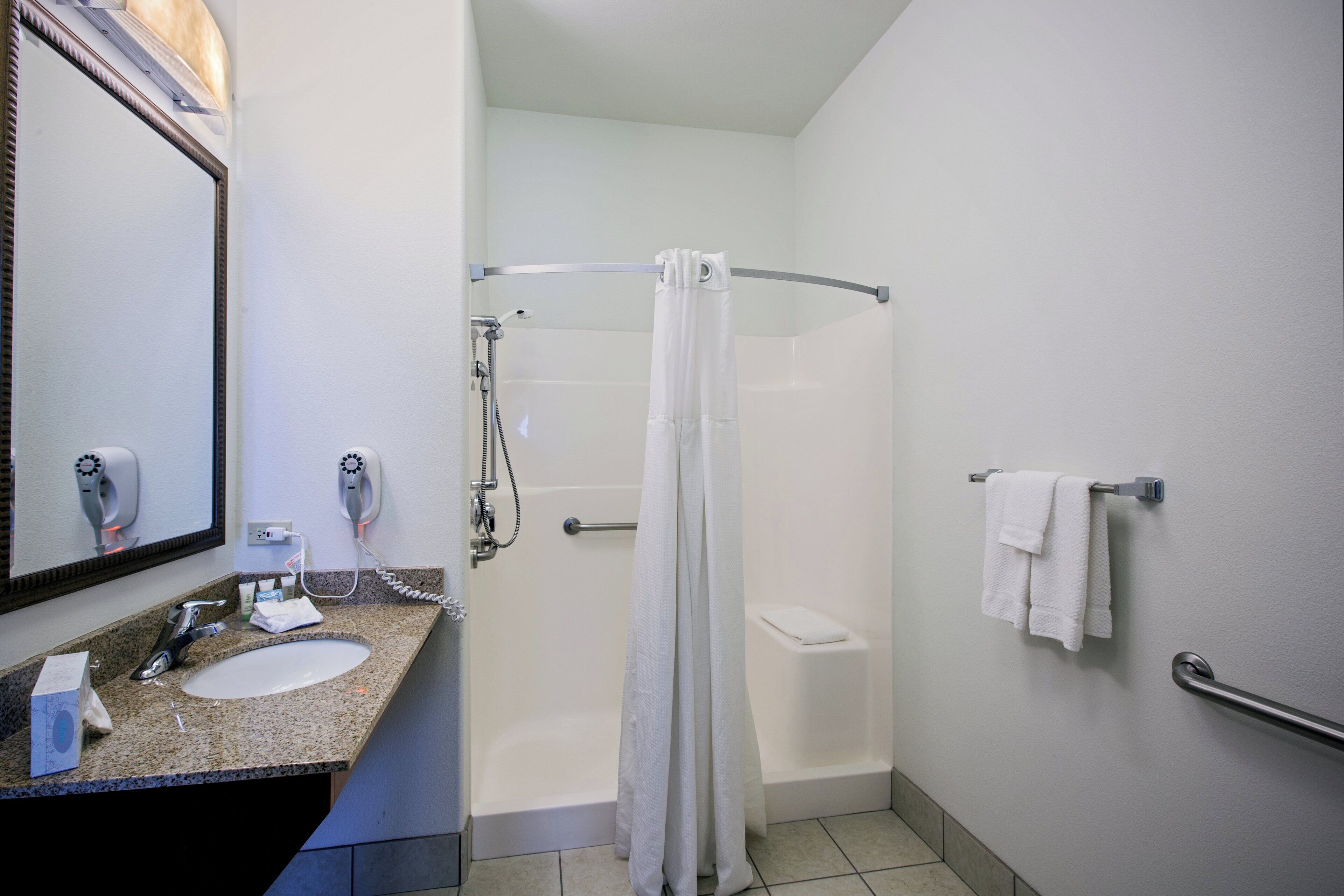 A bathroom with a granite countertop, a mirror, a hairdryer, a towel rack, and a shower with a curtain.