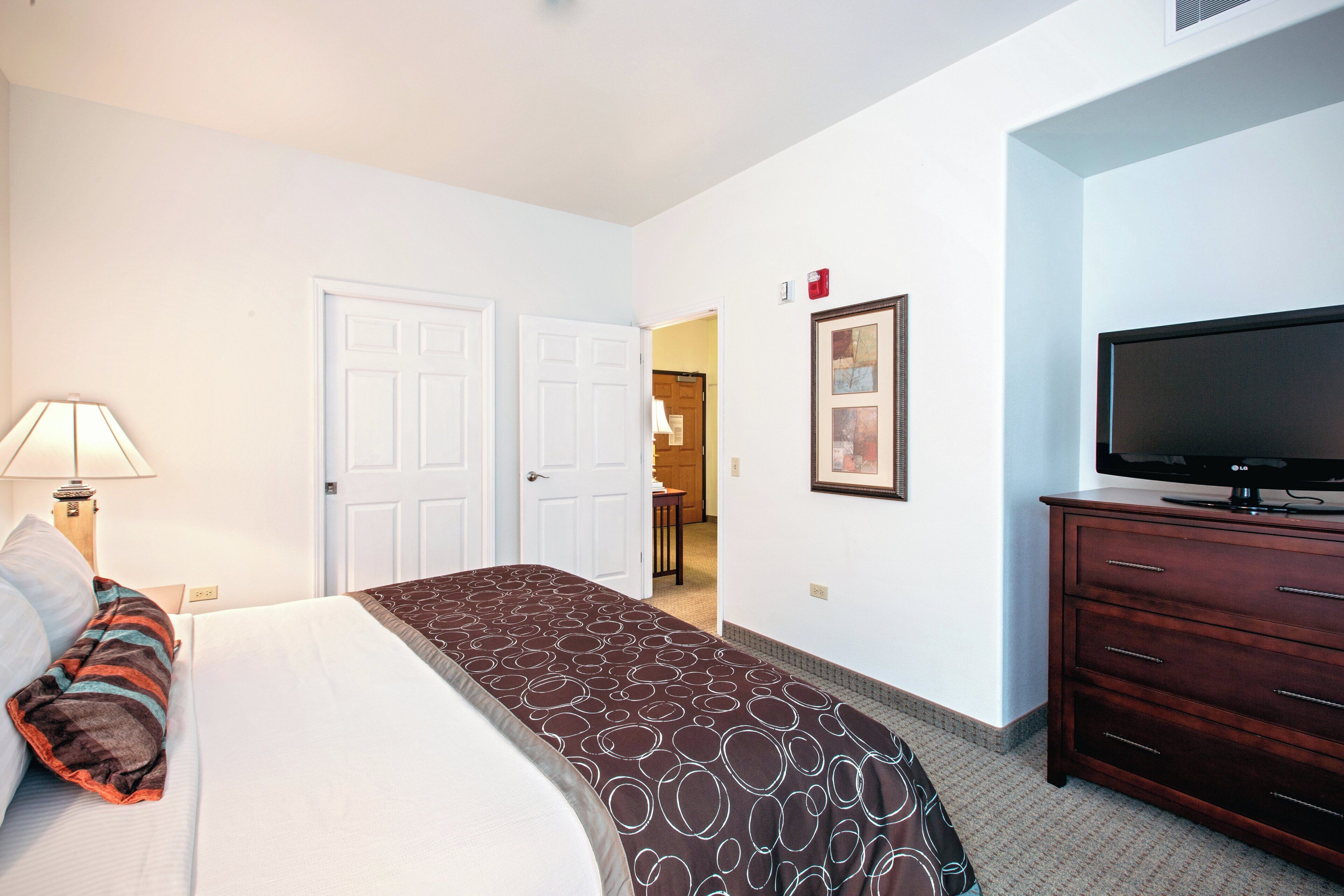 A hotel room with a bed, a TV, a dresser, and a door leading to another room.