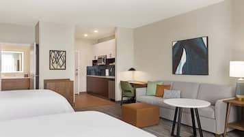 Habitación de hotel con dos camas, un sofá, una pequeña mesa, una lámpara, zona de cocina y obras de arte en las paredes.