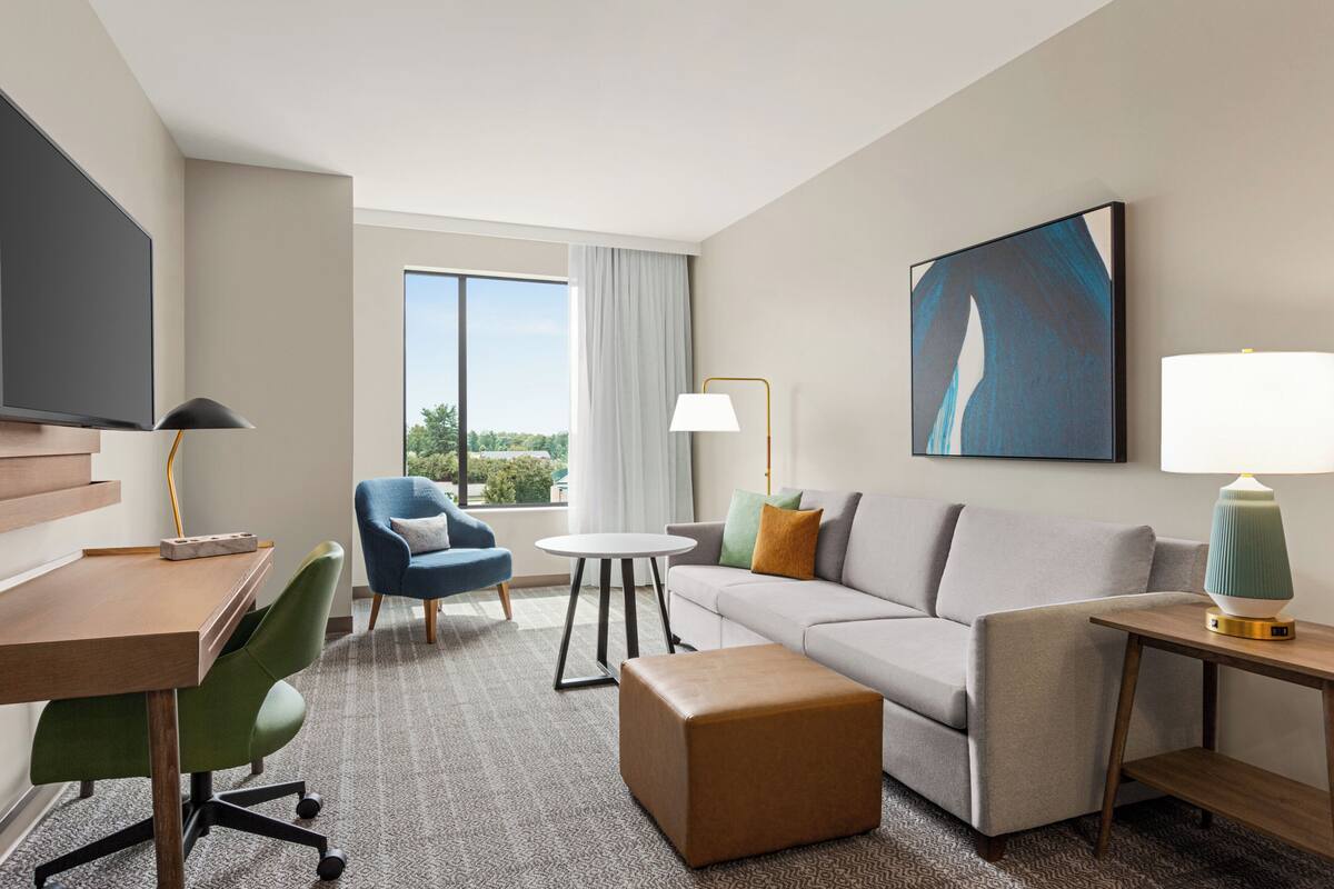 A hotel room with a flat-screen TV, a desk, a blue armchair, a gray sofa, a round coffee table, a lamp, and a painting on the wall.
