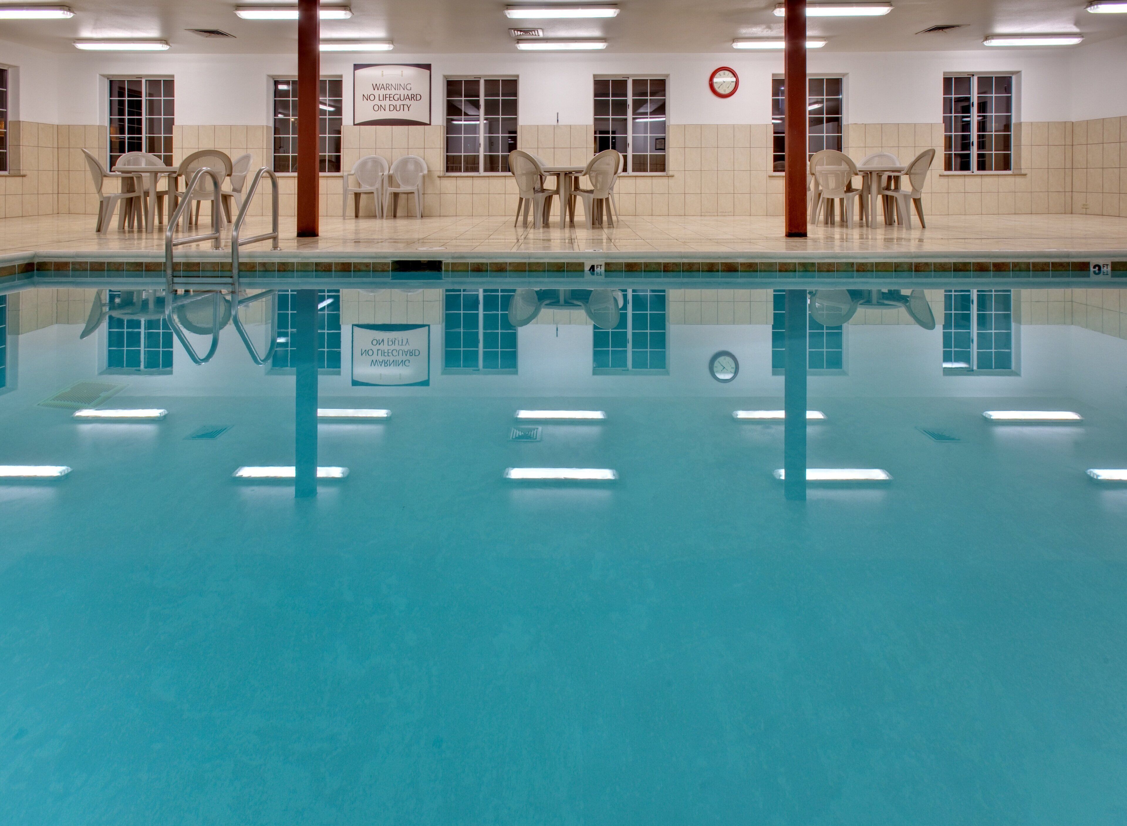 An indoor swimming pool with clear water, surrounded by chairs and tables, and signs indicating no lifeguard on duty.