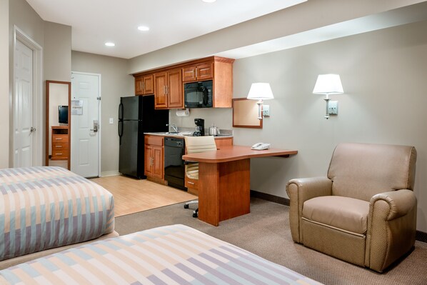 A compact hotel room with two beds, a kitchenette, a desk, and a chair.