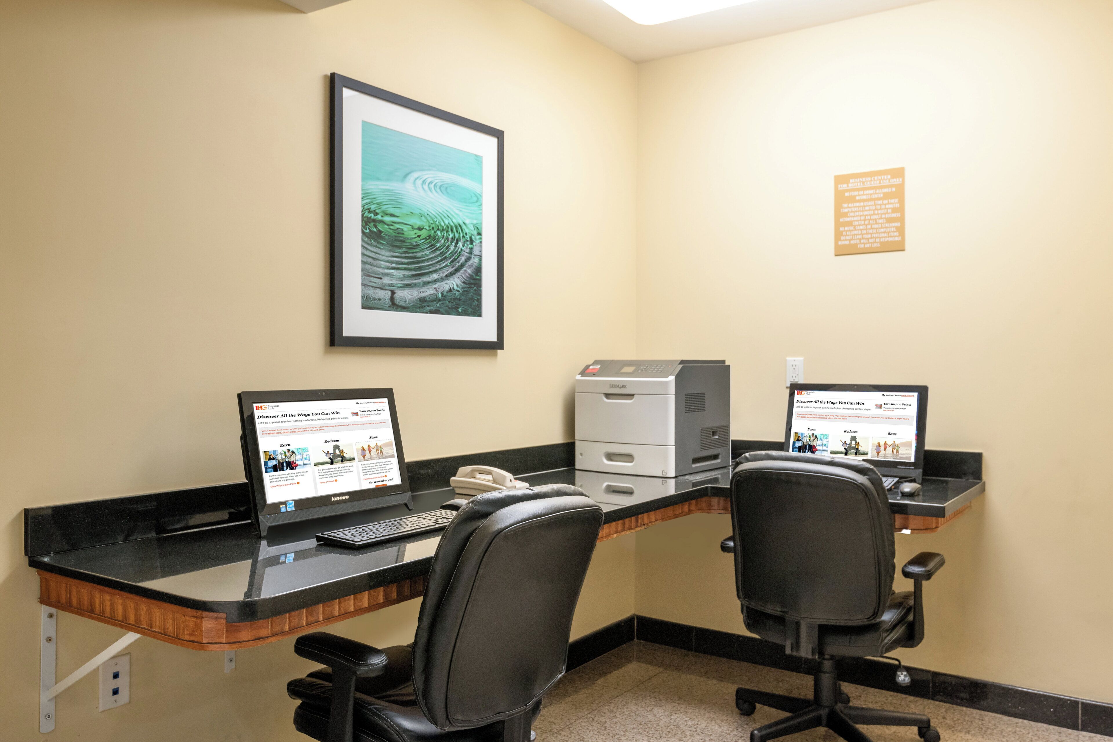 A workspace with two desks, each with a laptop, a phone, and a printer. There is a framed picture on the wall and a sign on the wall.