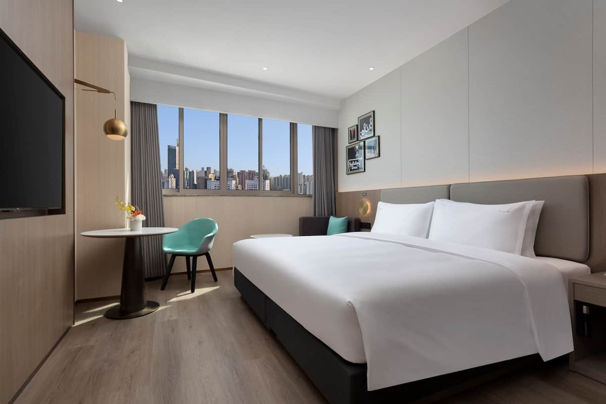 A modern hotel room with a large bed, a desk with a chair, a TV, and a city view.