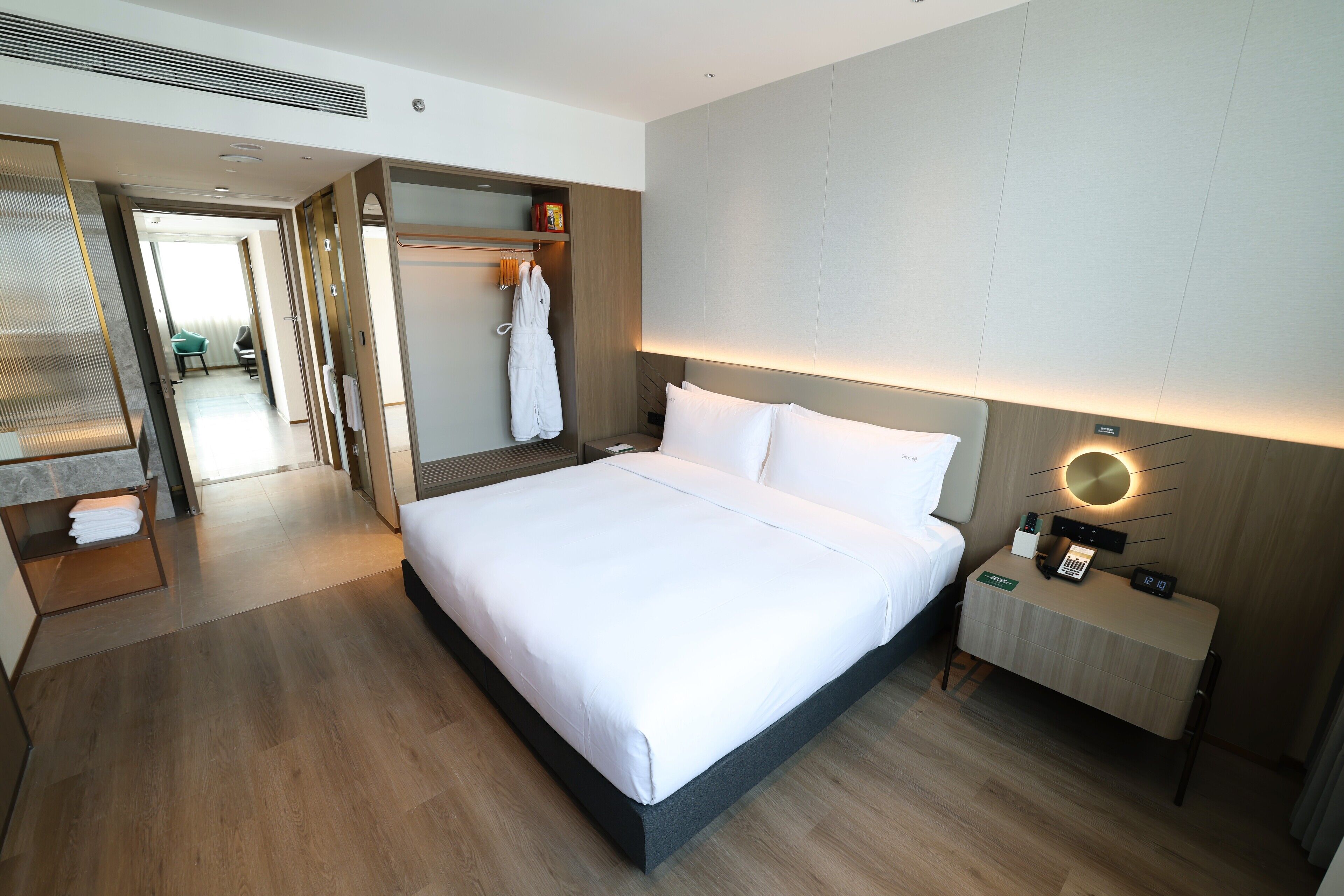 A modern hotel room with a large bed, a bedside table with a lamp, a wardrobe, and a bathroom visible in the background.