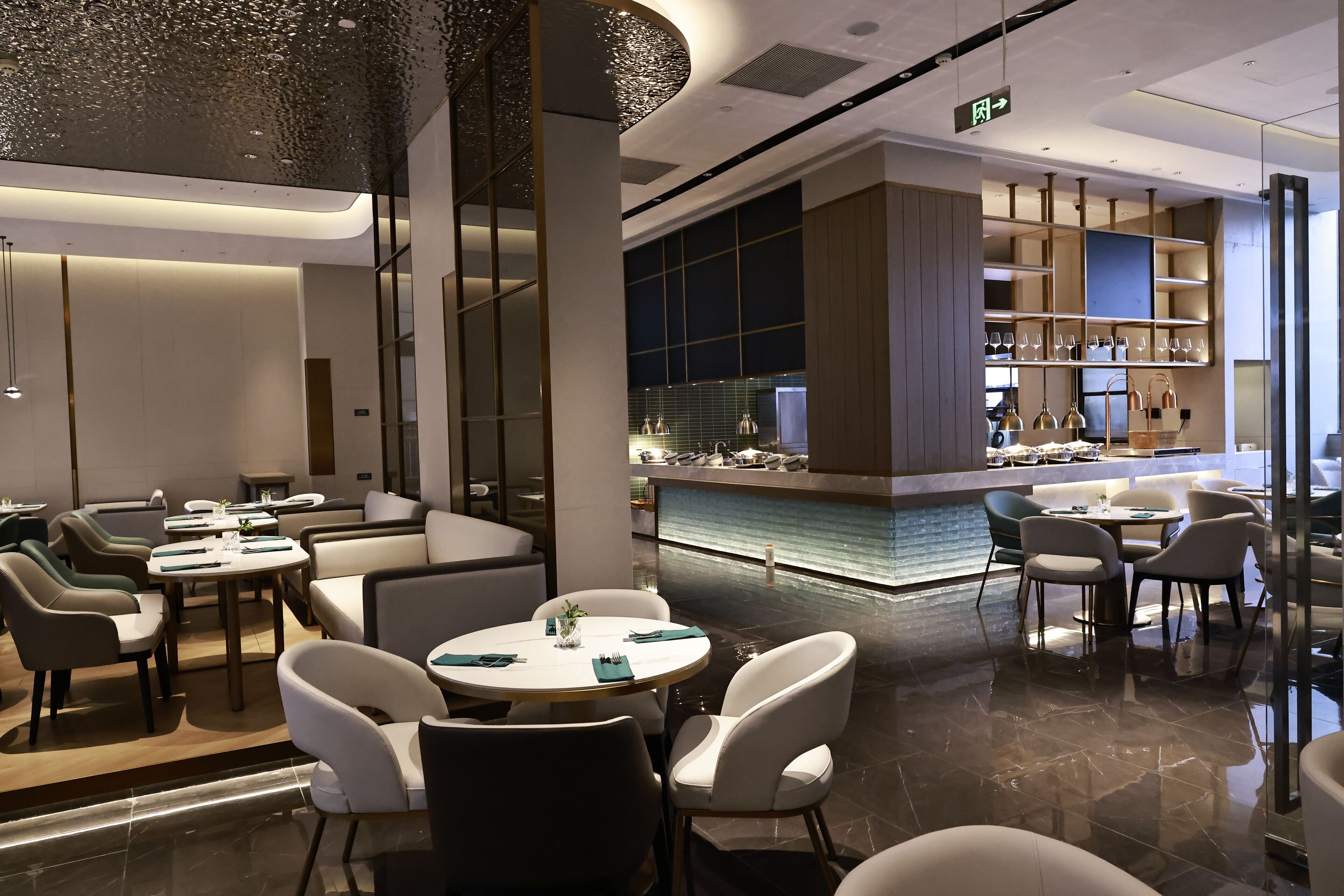 A modern restaurant with round tables and white chairs, a bar area with shelves of glasses, and a tiled floor.