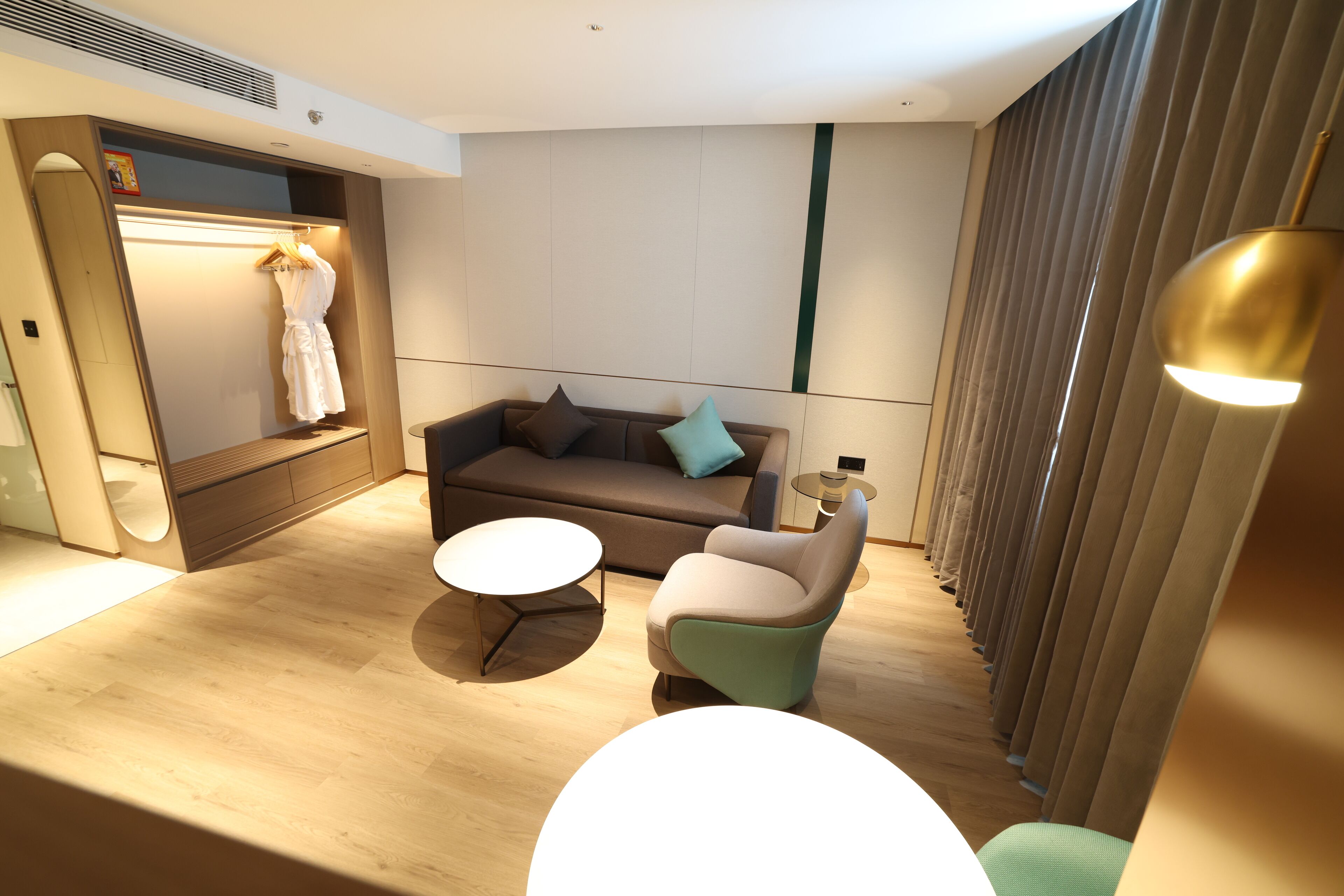 Una habitación de hotel moderna con piso de madera, un sofá marrón, una mesita redonda y una zona de estar con una silla verde.
