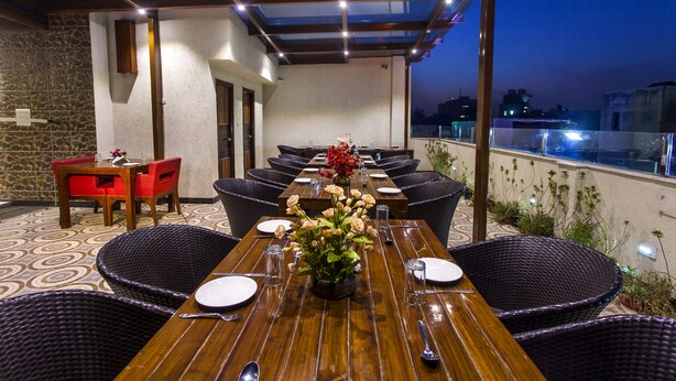 A rooftop dining area with a long wooden table set for six, surrounded by wicker chairs, and a smaller table with red chairs. - Hotel Sunstar Heritage (New Delhi)