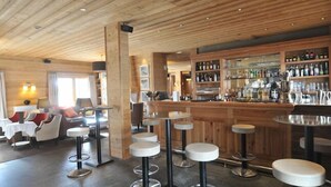 Una zona de bar con techo de madera y taburetes, una barra con botellas y un área de asientos con sillas y mesas.