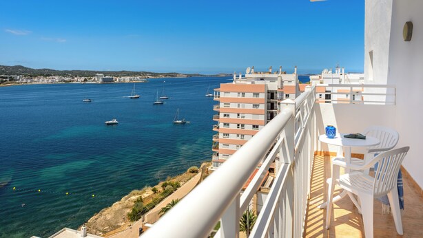 A balcony with a view of the sea, a table with chairs, and a bottle.
