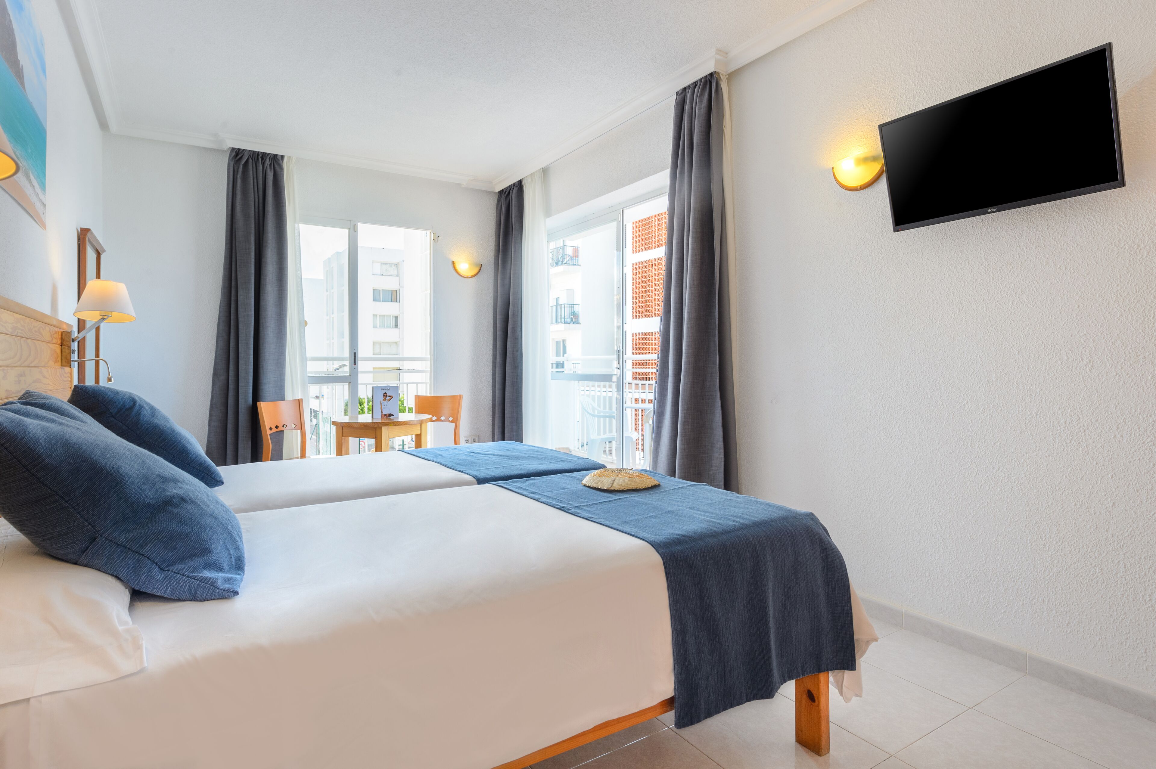 A hotel room with two beds, a TV mounted on the wall, a small table with a chair, and a balcony with curtains.