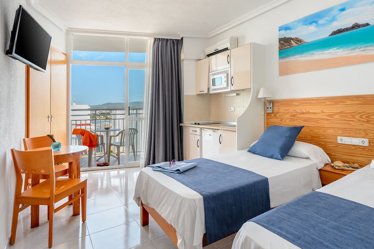 Quarto de hotel com duas camas, uma escrivaninha, uma cadeira, uma TV, varanda com vista e cozinha.