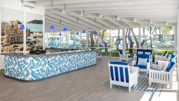 Amplio espacio interior con mostrador de recepción, mostrador de diseño azul y blanco, sillas blancas con cojines azules y vistas a una playa a través de amplias ventanas.