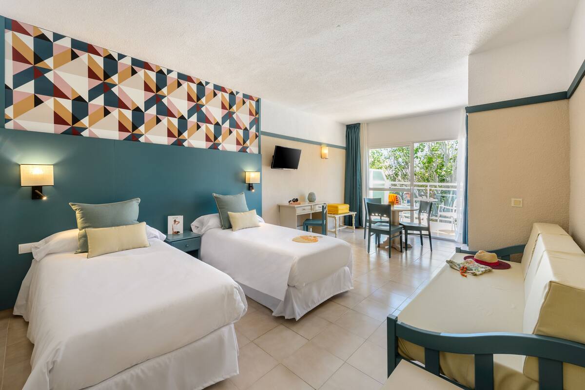 A hotel room with two beds, a desk, a chair, and a balcony with a table and chairs.