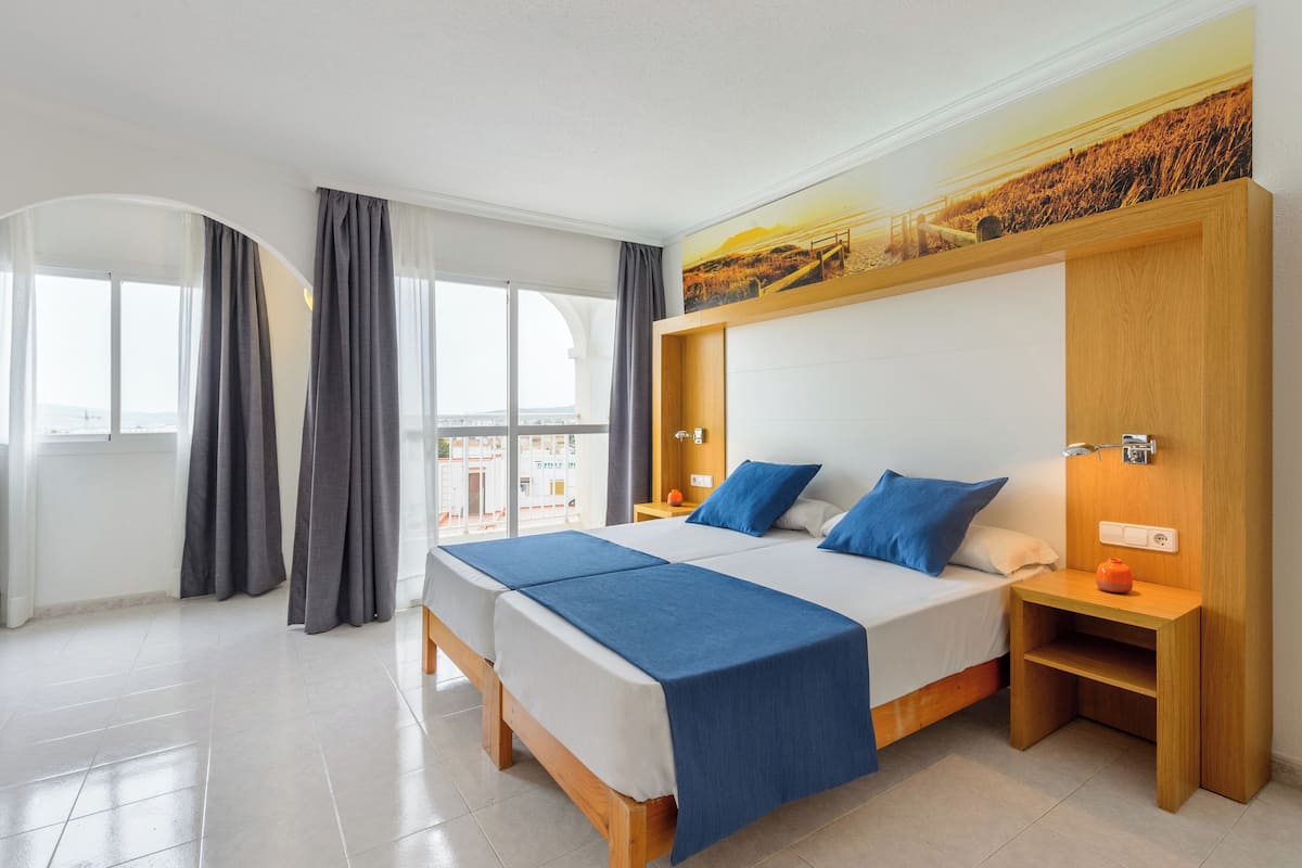A modern hotel room with a large bed, a wooden nightstand, a small table, and a view of the city through large windows.