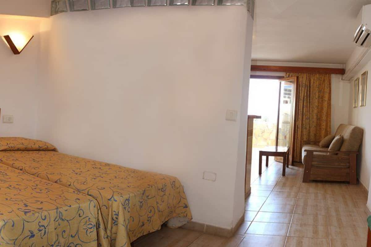 Una camera da letto con un letto, comodini, tenda da doccia, un water e un piccolo angolo salotto con una sedia e un tavolo.