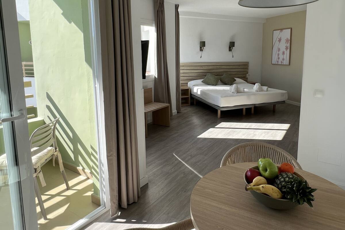 A hotel room with a bed, a bedside table with a fruit bowl, a chair, and a balcony with a chair.