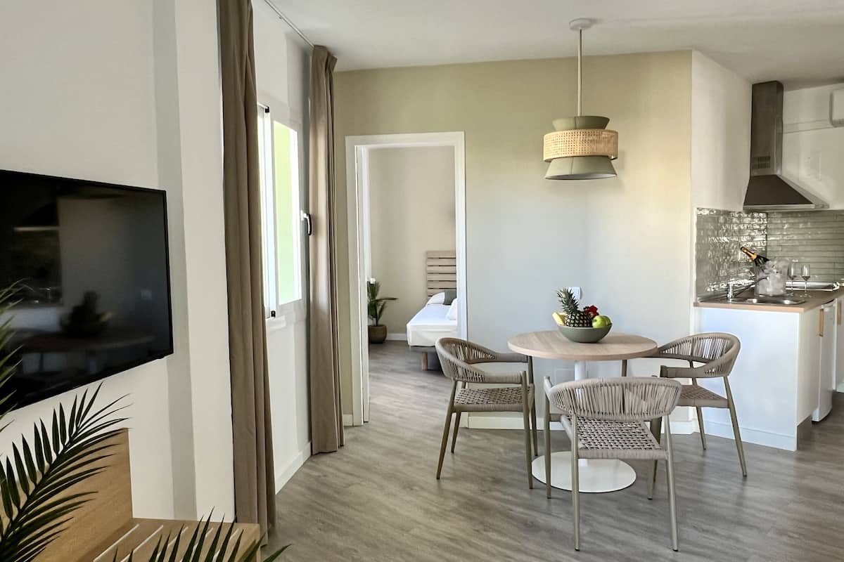 A modern living room with a flat-screen TV, a dining area with wicker chairs, and a kitchen with a built-in island.