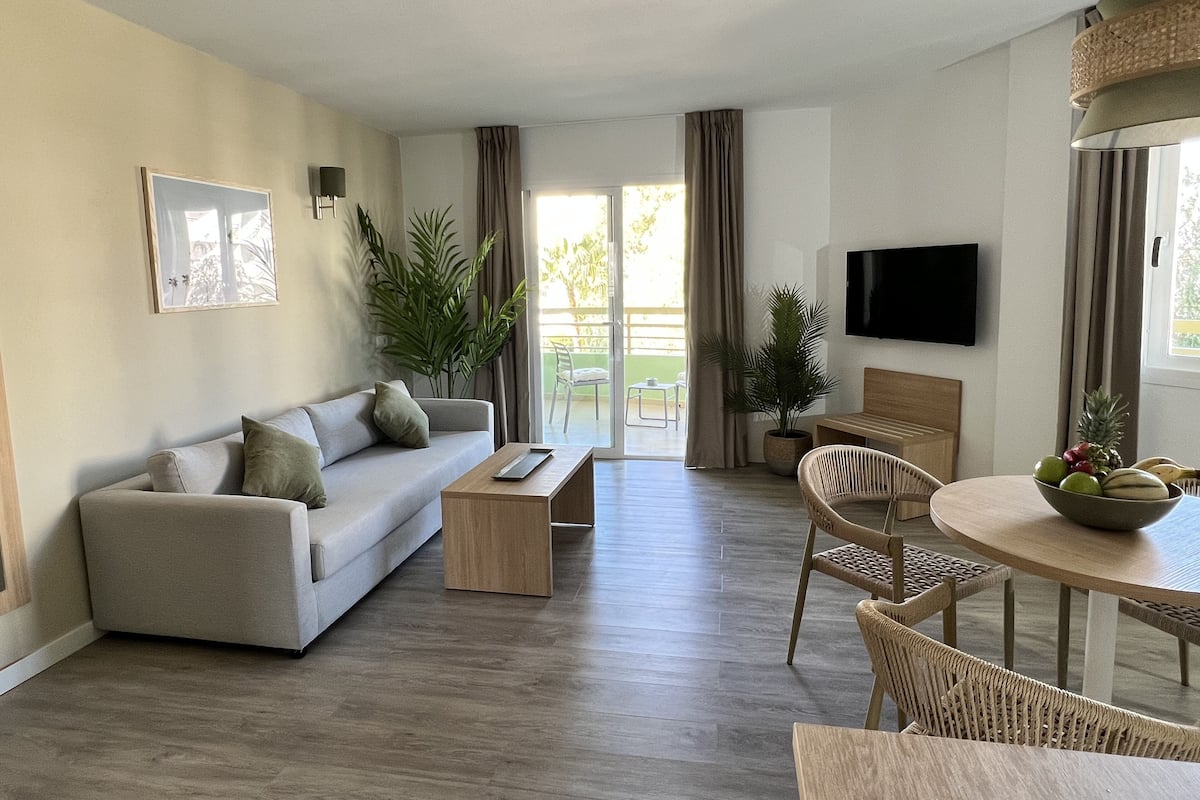A modern living room with a sofa, coffee table, and dining area. There is a TV mounted on the wall and a balcony visible through the glass doors.