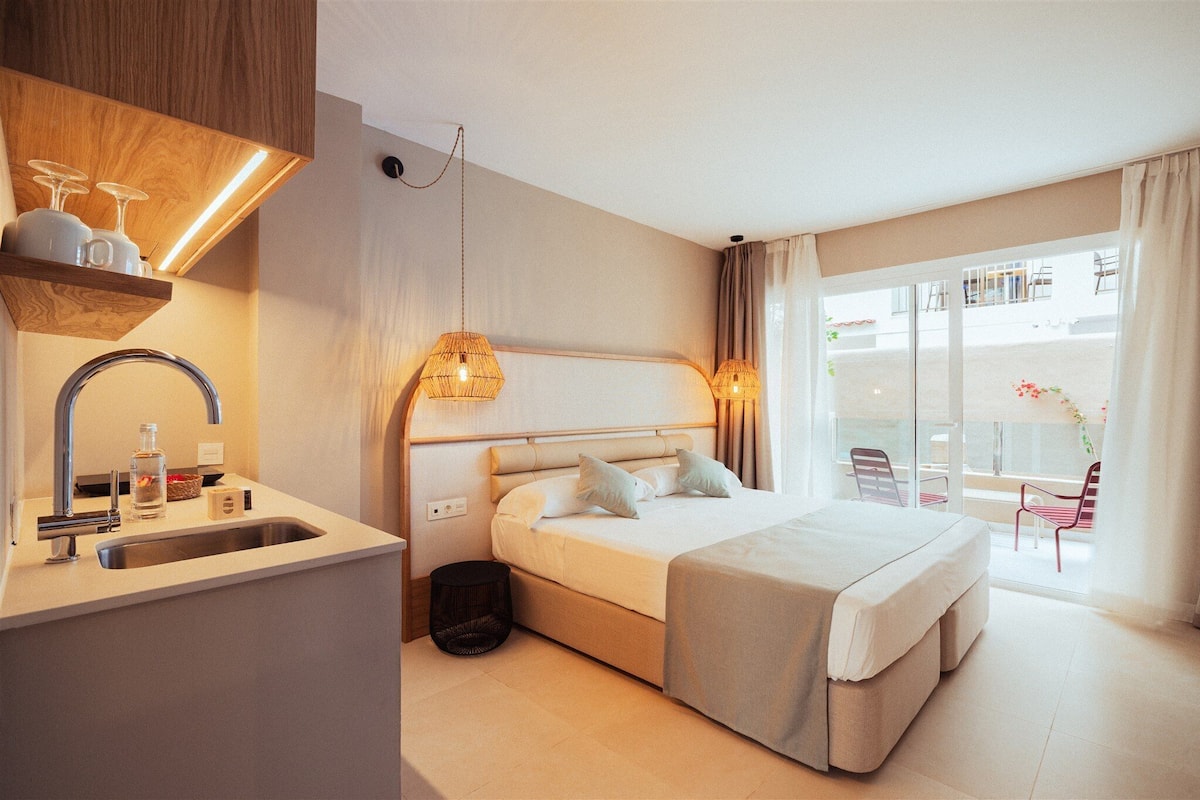 A modern hotel room with a large bed, a built-in sink, and a balcony with chairs.