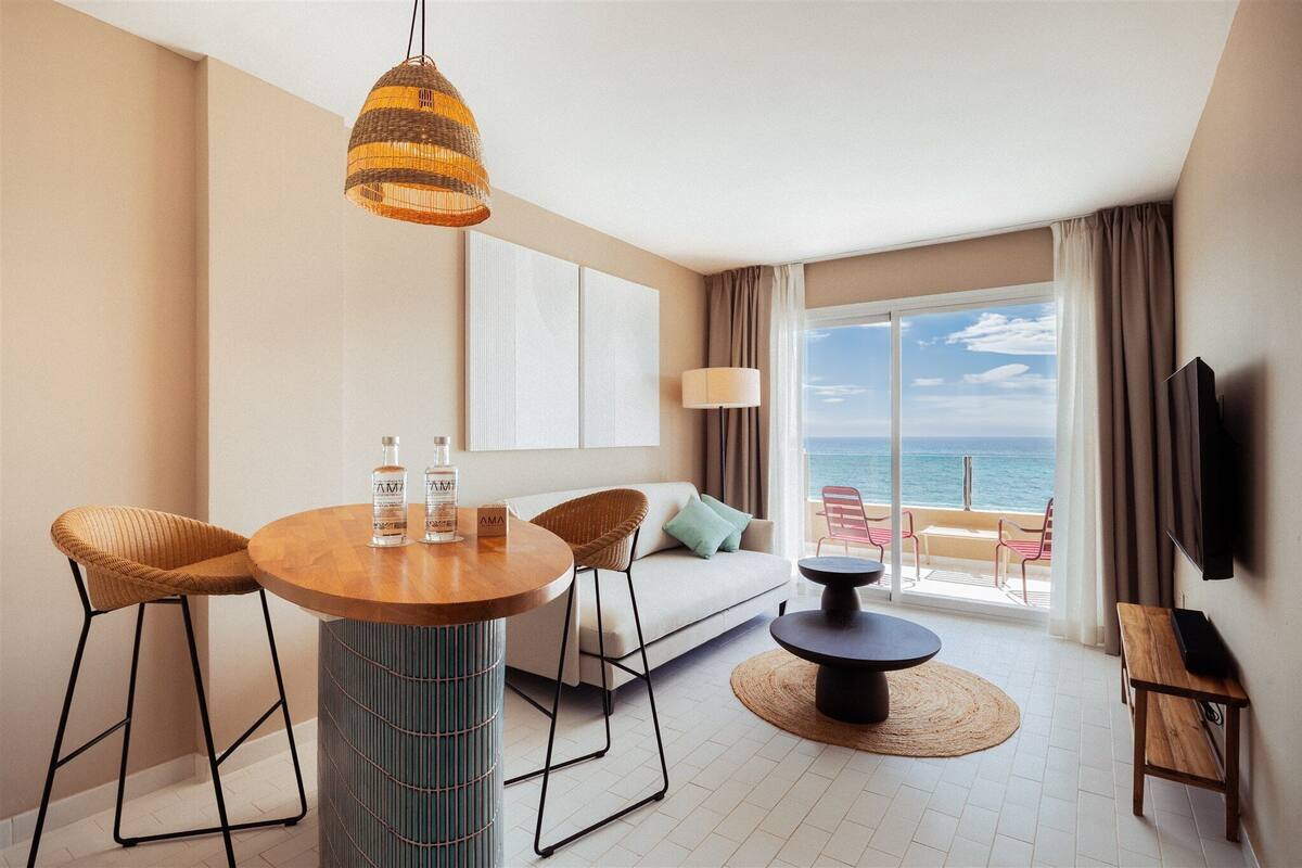 A modern hotel room with a round dining table, two bar stools, a sofa, a TV, and a large window with a view of the sea.