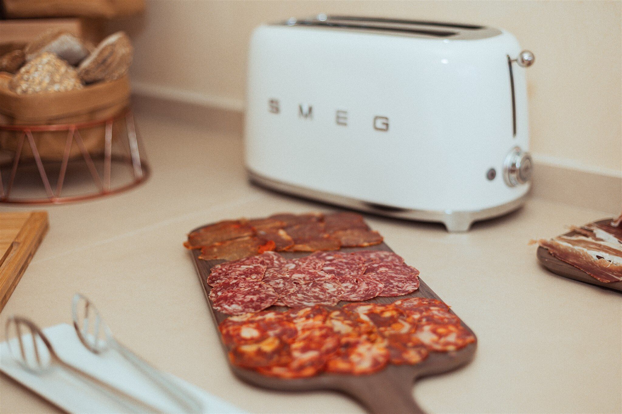 Una encimera de cocina con una tabla de madera que sostiene salami en rodajas, un tostador blanco y una cesta de pan.