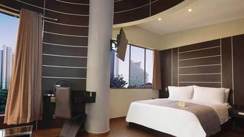A modern hotel room with a large bed, a flat-screen TV mounted on the wall, a desk with a chair, and a large window offering a city view.