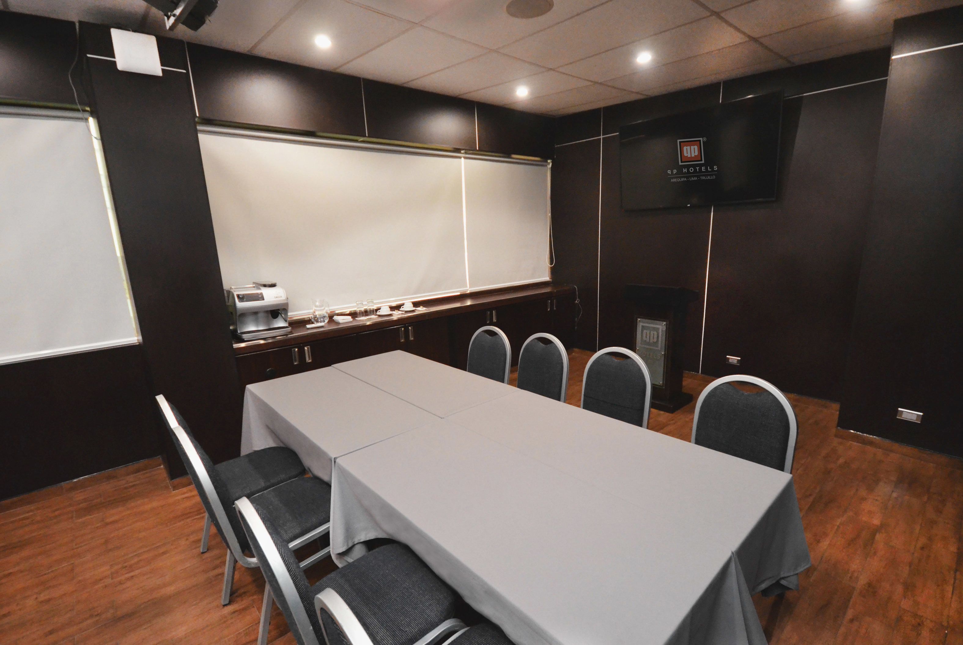 A conference room with a long table, chairs, a projector screen, a coffee machine, and a TV on the wall.