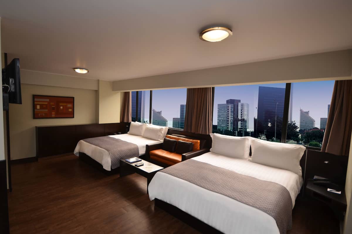 Ein Hotelzimmer mit zwei Betten, einem Sofa, einem Fernseher und einem großen Fenster mit Blick auf die Stadt.