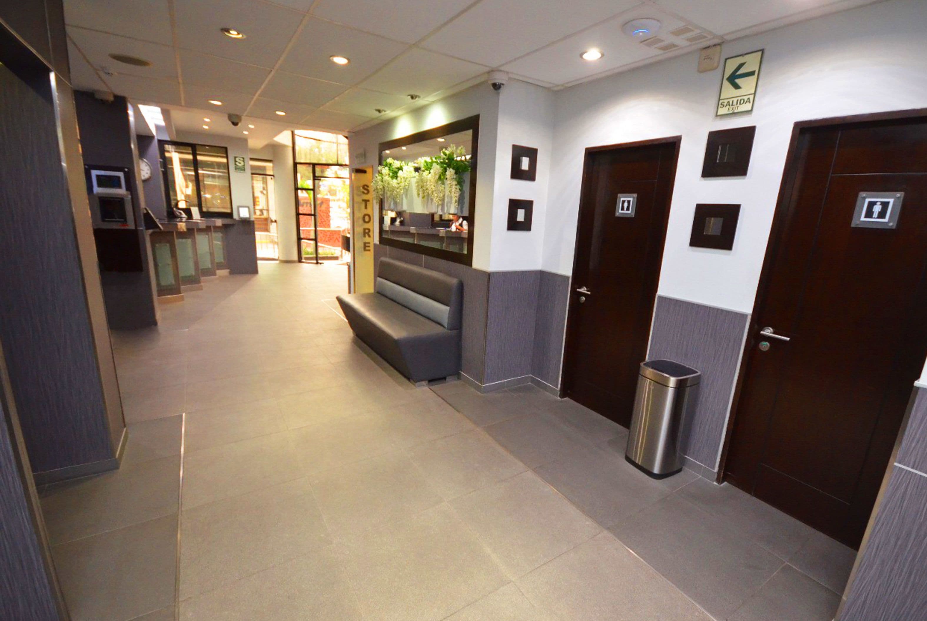 A hotel lobby with a reception desk, a bench, a trash bin, and doors marked with an exit sign.